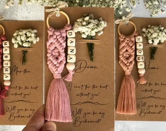 May include: Three macrame keychains in shades of pink, each attached to a brown card with the text 'I can't tie the knot without you! Will you be my Bridesmaid?' and a small bouquet of dried white baby's breath flowers attached to each card.