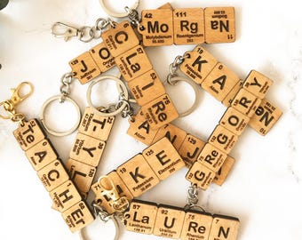 May include: A collection of keychains made from light-colored wood squares, each featuring black printed letters and numbers resembling periodic table elements. The squares are linked together to spell names like Morgan, Claire, Kay, Teacher, and Lauren.