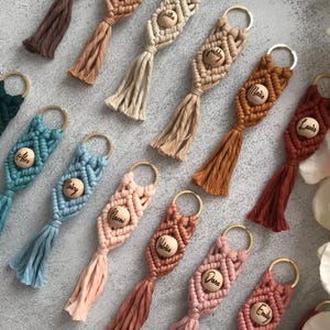 Puede incluir: Una colección de llaveros de macramé en varios colores, cada uno con una cuenta de madera central grabada con un nombre y una anilla de metal dorado. Los diseños de cordón anudado terminan en una borla, en tonos marrones, azules, rosas y rojos.