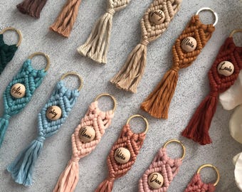 May include: A collection of macrame keychains in various colors, each featuring a central wooden bead engraved with a name and a gold-toned metal ring. The knotted cord designs end in a tassel, showcasing shades of brown, blue, pink, and red.