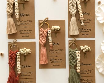 May include: Five macrame keychains in beige, dusty pink, terracotta, and sage green, each attached to a gold ring and a brown card. The cards feature dried white flowers, names in wooden beads, and the text: I can't say I Do without you!