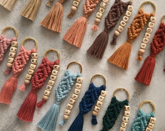 May include: A collection of macrame keychains in various colors including beige, pink, brown, orange, blue, and green. Each keychain features a gold-tone metal ring, a knotted cord design, and a wooden bead strand with a name printed in black letters.