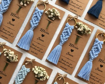 May include: Light blue macrame keychains with gold-tone rings attached to rectangular kraft paper cards. Each card features the printed names Maria and Jason, a heart symbol, and the text Thank you for celebrating with us! 02.05.2022, with dried flowers.