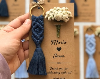 May include: A navy blue macrame keychain with a tassel attached to a gold ring, mounted on a rectangular kraft paper card. The card features a small bouquet of white baby's breath flowers and text reading Maria, Jason, Thank you for celebrating with us! 02.05.2022.
