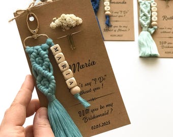 May include: A teal macrame keychain with a gold-tone lobster clasp and a wooden bead strand spelling MARIA, attached to a brown card featuring the text Will you be my Bridesmaid? and a small dried flower sprig, with other similar keychains visible.