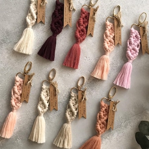 May include: A collection of nine macrame keychains in various colors including cream, burgundy, dusty rose, peach, and light pink. Each keychain features a gold-tone metal clasp and a wooden tag engraved with the text "Julia & Ethan September 20, 2022".