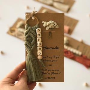 Peut inclure: Un porte-clés en macramé vert sauge avec un anneau doré et des perles en bois avec les lettres AMANDA, fixé sur une carte en papier kraft rectangulaire ornée d'un brin de fleurs séchées blanches et du texte Will you be my Maid of Honor?
