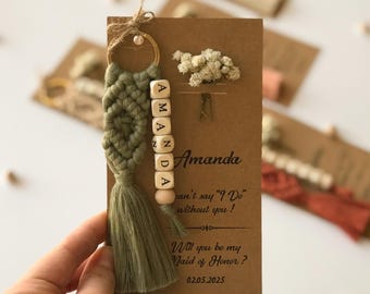 May include: A sage green macrame keychain with a gold ring and wooden letter beads spelling AMANDA, attached to a rectangular kraft paper card featuring a dried white flower sprig and printed text including Will you be my Maid of Honor?