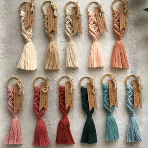 Pode incluir: Onze porta-chaves de macramé em várias cores, incluindo creme, bege, rosa, terracota, verde escuro e azul. Cada porta-chaves possui um anel de metal dourado, um design de corda atada, uma borla e uma etiqueta de papel kraft com texto.