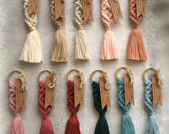 May include: Eleven macrame keychains in various colors including cream, tan, pink, terracotta, dark green, and blue. Each keychain features a gold-tone metal ring, a knotted cord design, a tassel, and a rectangular kraft paper tag with text.
