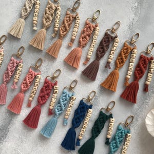 Op de afbeelding: Een collectie macramé sleutelhangers in diverse kleuren zoals beige, roze, terracotta, bruin, blauw en groen. Elke sleutelhanger is voorzien van een geknoopt koordontwerp, een goudkleurige metalen sluiting, een kwastje en houten kralen met namen.