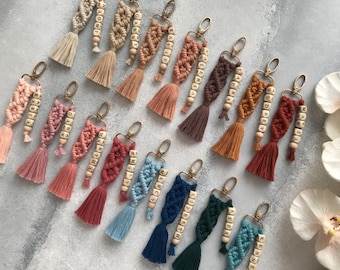 May include: A collection of macrame keychains in various colors including beige, pink, terracotta, brown, blue, and green. Each keychain features a knotted cord design, a gold-toned metal clasp, a tassel, and wooden beads spelling out names.