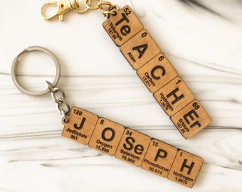May include: Two wooden keychains featuring square tiles engraved with periodic table elements. One keychain spells TEACHER using Te, A, C, H, Er, and the other spells JOSEPH using J, O, Se, P, H. Each tile includes atomic numbers and weights.