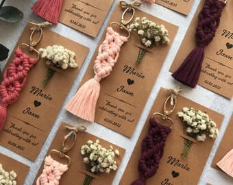 May include: Macrame keychains in shades of pink, rose, and deep purple, each attached to a gold-tone metal clasp. The keychains are mounted on rectangular kraft paper cards printed with the names Maria and Jason and the date 02.05.2022.