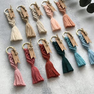 Op de afbeelding: Een collectie van elf macramé sleutelhangers in diverse kleuren, waaronder crème, beige, roze, terracotta, donkergroen en blauw. Elke sleutelhanger heeft een gouden metalen ring en een bruin papieren label met de tekst Kinda & Ethan September 30, 2022.