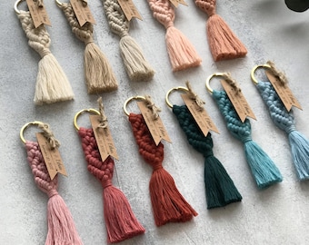 May include: A collection of eleven macrame keychains in various colors including cream, beige, pink, terracotta, dark green, and blue. Each keychain features a gold metal ring and a small brown paper tag printed with the text Kinda & Ethan September 30, 2022.