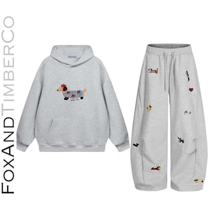 Può includere: Un completo composto da una felpa con cappuccio e pantaloni della tuta a gamba larga in grigio chiaro. La felpa presenta un bassotto ricamato con basco e maglione. I pantaloni sono decorati con diversi piccoli bassotti e cuori ricamati.
