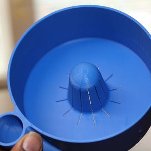 May include: A bright blue, circular, 3D-printed plastic tool featuring a central, cone-shaped reamer with vertical slits and a small, cylindrical side attachment. The object has a smooth, matte finish and a uniform, solid blue color throughout.