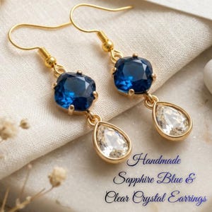 Puede incluir: Un par de pendientes colgantes dorados con un cristal azul zafiro de corte cuadrado unido a un cristal transparente en forma de lágrima. El texto Handmade Sapphire Blue & Clear Crystal Earrings aparece en azul en la parte inferior derecha.