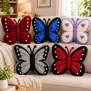May include: Five butterfly-shaped crochet pillows in various colors. The top row features a deep red pillow with gold beads, a blue pillow with white beads, and a cream pillow with purple hearts. The bottom row has a gray pillow with white beads and a bright red pillow with gold beads.
