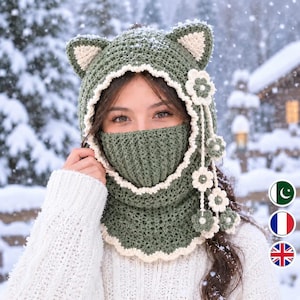 Puede incluir: Un pasamontañas de ganchillo verde oliva con orejas de gato con centros color crema, un borde festoneado color crema y una tira colgante de flores de ganchillo color crema y verde oliva. La capucha cubre la cabeza y el cuello.