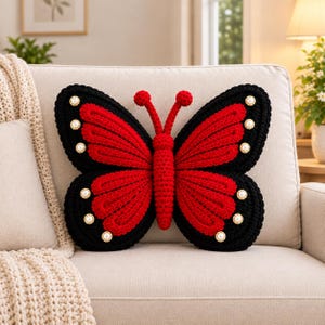 May include: A butterfly-shaped crochet pillow featuring a vibrant red center and wings with black borders. The wings are decorated with small, round, white pearl-like beads arranged along the edges. The pillow has two red antennae on top.