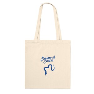 May include: A beige canvas tote bag featuring two long shoulder straps and a centred graphic print in blue. The design consists of the text "Beware of Snakes" written in a cursive script font positioned above a simple blue silhouette of a snake.