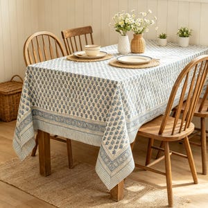 May include: A rectangular tablecloth featuring a blue and white block-print pattern with a repeating floral motif, a wide decorative border, and striped edges. The fabric drapes over a wooden dining table set with woven placemats and dishes.