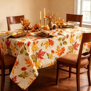 May include: A rectangular dining table covered with a cream-colored tablecloth featuring a vibrant pattern of autumn leaves in shades of red, orange, yellow, and green. The table is set with amber-colored glassware, candles, and small pumpkins.