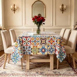 May include: A rectangular dining table covered with a tablecloth featuring a vibrant, multi-colored tile pattern in shades of red, blue, yellow, and white. A clear glass vase filled with red roses sits in the center of the table.