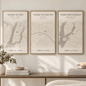 May include: Three vertical framed wall art prints featuring beige map backgrounds with dark gray street lines. Each print displays text: Where We Met New York, Where We Said Yes Paris, and Where Forever Began Brooklyn, with dates and names Emma & Liam.