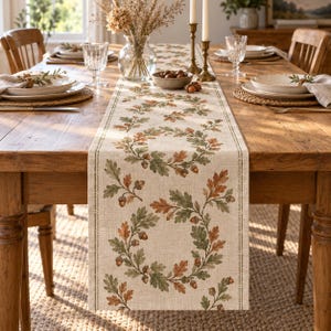 May include: A beige linen table runner featuring a repeating pattern of circular wreaths made of green and orange oak leaves and brown acorns. The runner has a thin vertical border line on each side and is centered on a wooden dining table.