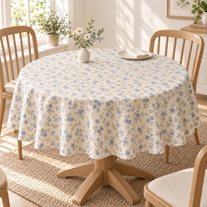 May include: A round tablecloth featuring a cream-coloured base with a repeating pattern of small blue flowers and green leaves. The fabric drapes over a circular wooden table, which is surrounded by wooden chairs in a bright, sunlit room setting.