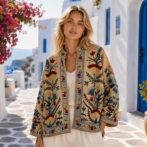 Könnte beinhalten: Eine beige, vorne offene Jacke mit aufwendiger Blumenstickerei in Rot-, Blau- und Gelbtönen. Das Kleidungsstück hat einen Stehkragen, lange Ärmel und einen strukturierten Saum und wird über einem schlichten weißen Oberteil und weißer Hose getragen.