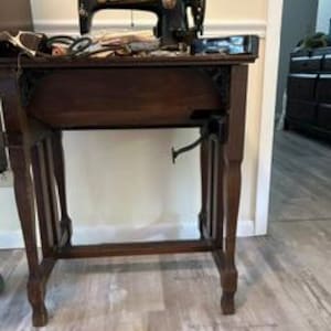 May include: A vintage black sewing machine with gold lettering mounted on a dark brown wooden table. The machine features a hand wheel on the right side and a metal treadle mechanism underneath. Various sewing tools and fabric scraps sit on the table.