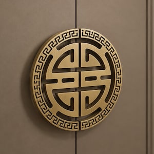 May include: A circular brass cabinet handle mounted on a gray door. The handle features a symmetrical cutout design with a geometric Greek key border pattern around the outer edge, split into two halves that meet in the center of the door.