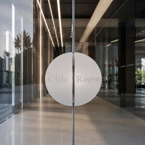 May include: A circular white door handle split into two halves, mounted on a glass door. The left half features the embossed text Chic and the right half features the embossed text Room. The handle is positioned centrally on the glass surface.