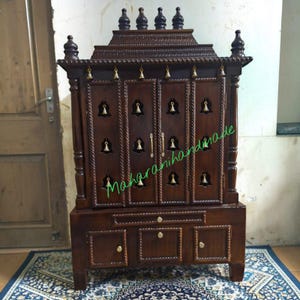 May include: A dark brown wooden home temple featuring four doors with brass bell accents, a tiered top with four finials, and a base with three drawers. The structure has carved rope-style borders and gold-toned hardware on the drawers.