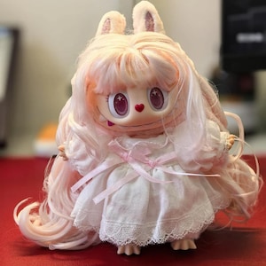 May include: A doll with long, pale pink hair and rabbit-like ears, featuring large purple eyes with star-shaped highlights and a small red heart-shaped nose. The doll wears a white dress with lace trim and a light pink ribbon tied at the waist.