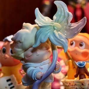 May include: A collectible figurine of a character with closed eyes and light-colored hair, wearing a pale blue hooded jacket. The character holds a large, stylized flower with petals transitioning from pale blue to soft pink, mounted on a clear base.
