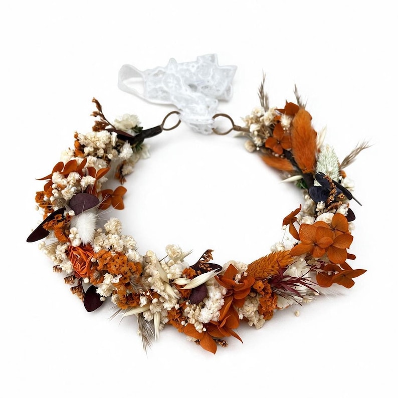 May include: A circular floral crown featuring a mix of dried white baby's breath, burnt orange hydrangea petals, rust-colored grasses, and dark brown leaves. The arrangement is secured with a white lace ribbon tied at the back with metal rings.