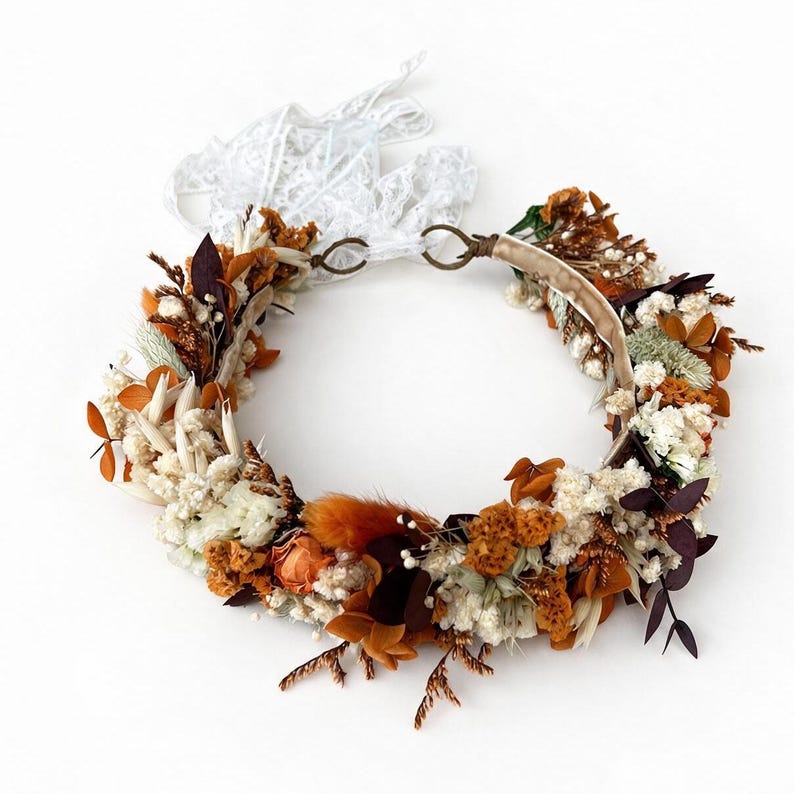 May include: A circular floral crown featuring a mix of dried white, burnt orange, and deep brown foliage. The arrangement includes small white flowers, orange-toned grasses, and dark leaves, secured with a beige band and white lace ribbons.