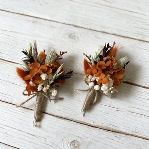 May include: Two boutonnieres featuring dried flowers in shades of burnt orange, cream, and dark brown. Each arrangement includes preserved hydrangea, fluffy bunny tails, and small white filler flowers, with stems wrapped in natural jute twine.
