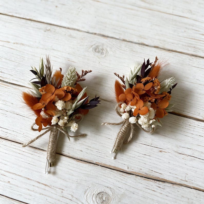May include: Two boutonnieres featuring dried flowers in shades of burnt orange, cream, and dark brown. Each arrangement includes preserved hydrangea, fluffy bunny tails, and small white filler flowers, with stems wrapped in natural jute twine.