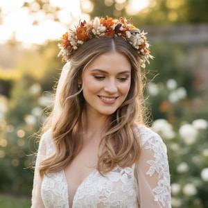 May include: A floral crown featuring dried flowers in shades of burnt orange, cream, and brown, worn atop long wavy hair. The headpiece is composed of small blossoms, grasses, and foliage arranged in a circular band, resting above a white lace dress.