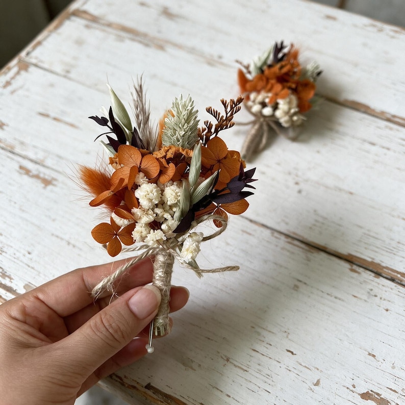 May include: A boutonniere featuring a collection of dried flowers in shades of burnt orange, cream, and dark brown. The arrangement includes small hydrangea petals, fluffy grasses, and sprigs, all bound together with twine around a wrapped stem.