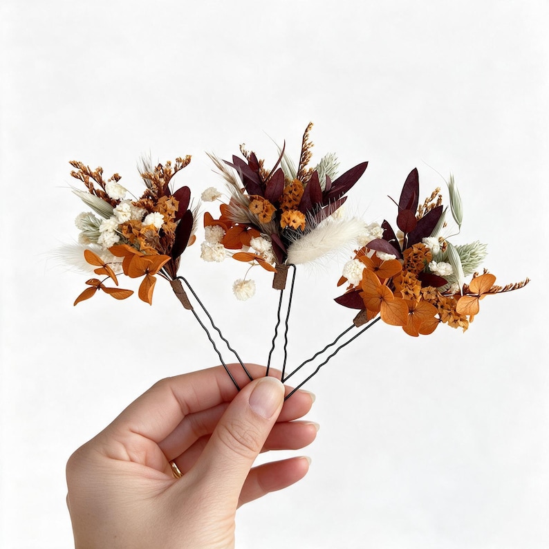 May include: Three hair pins featuring arrangements of dried flowers and foliage in shades of deep burgundy, burnt orange, white, and sage green. Each pin is constructed with a dark metal U-shaped base, topped with a cluster of preserved botanical elements.