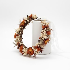 May include: A circular floral crown composed of dried orange flowers, white baby's breath, and various grasses. The arrangement features a rustic twig base and includes a long, sheer white ribbon attached to one side for tying around the head.