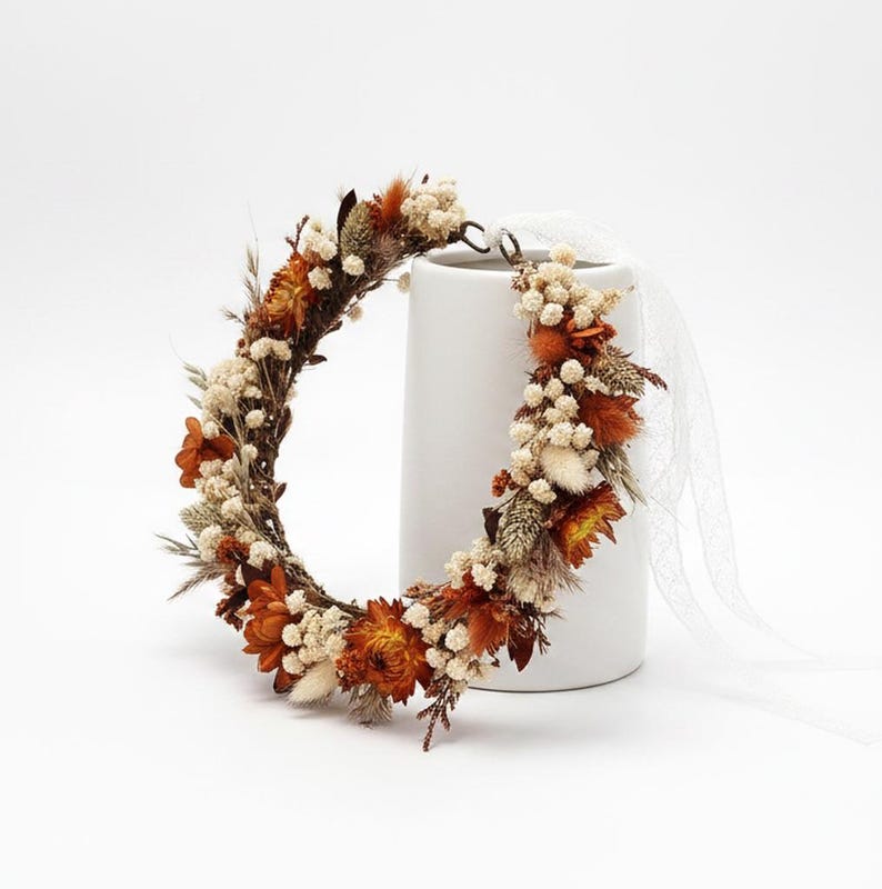 May include: A circular floral crown composed of dried orange flowers, white baby's breath, and various grasses. The arrangement features a rustic twig base and includes a long, sheer white ribbon attached to one side for tying around the head.