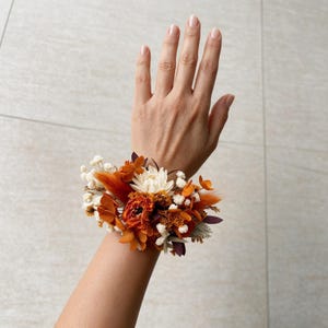 May include: A wrist corsage featuring a cluster of dried flowers in shades of burnt orange, deep rust, and cream. The arrangement includes small white baby's breath, textured orange petals, and dark maroon accents, secured together as a floral wrist accessory.